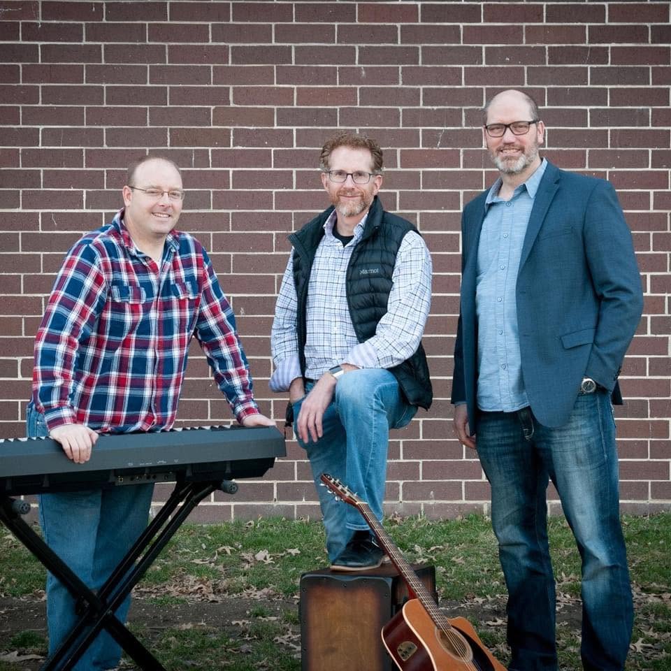 The three members of Looking4Luke standing together outdoors with a guitar, keyboard, and cajon